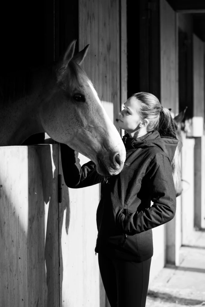 Photo noir et blanc de la cavalière Liv Piris portant la veste Mélanie Bleu Marine d'égérie élite rider ainsi que le pantalon d'équitation Nina Bleu Cobalt de la marque égérie élite rider posant avec un cheval blanc